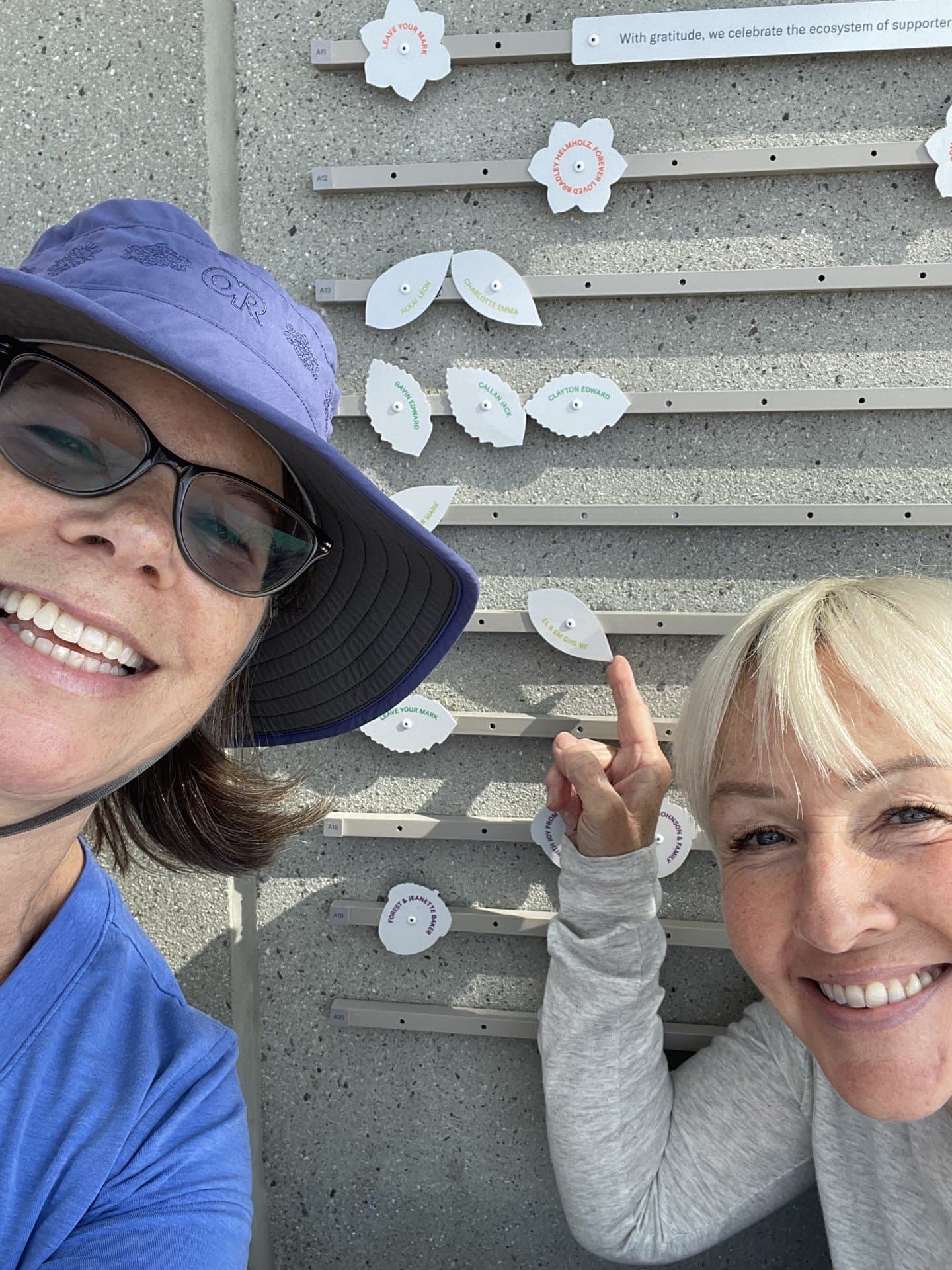 Two smiling women pose while pointing to their botanical token on the Community Wall at Overlook Walk.