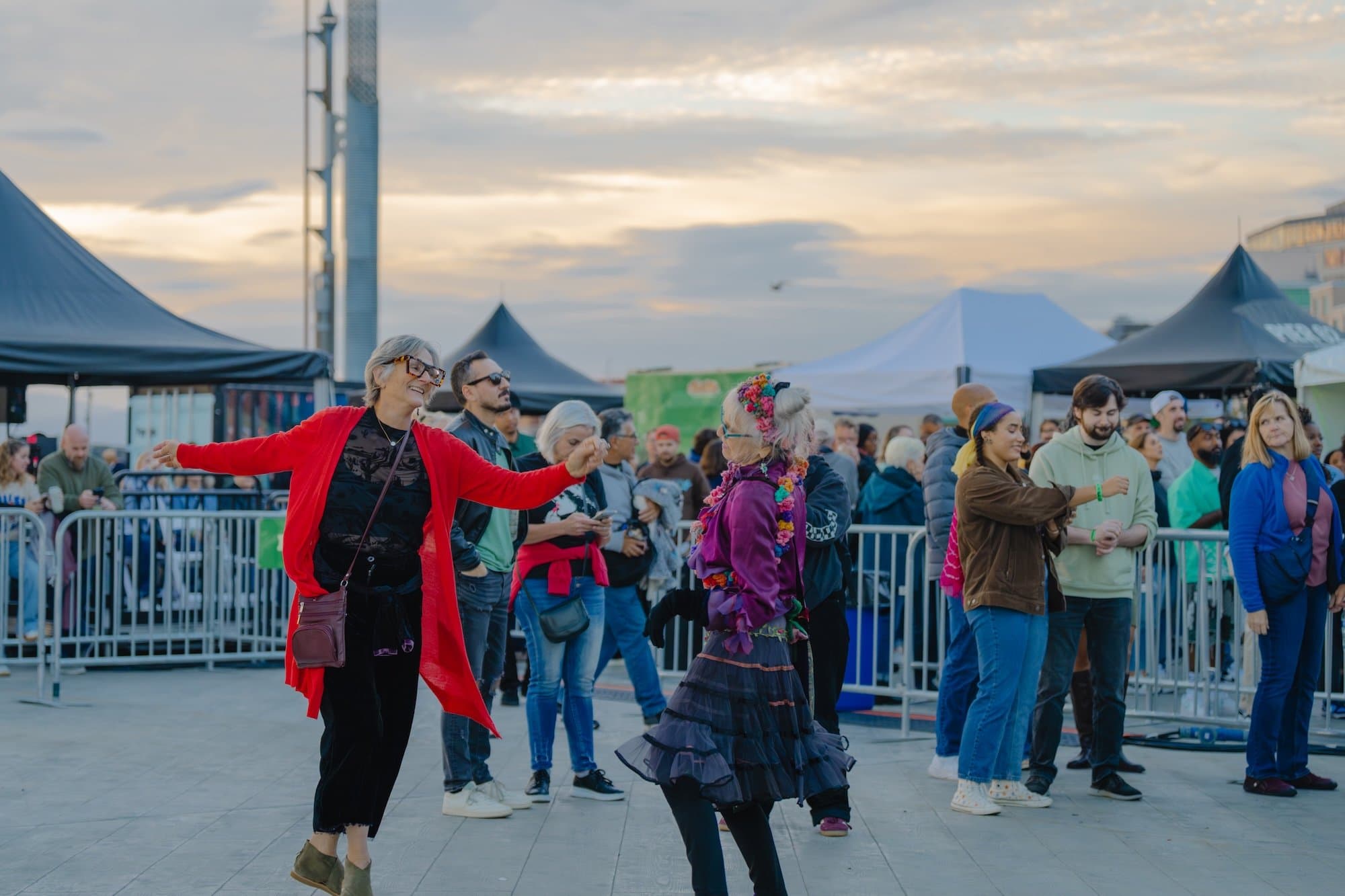 A person in a long red sweater dances with their arms outstretched at a concert on Pier 62, with groups of people and tents behind them.